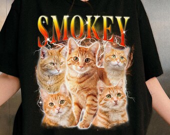 May include: Black t-shirt featuring a graphic of six orange tabby cats with the word "SMOKEY" in a yellow and red font. The cats are arranged in a group, with lightning bolt effects around them. The shirt is a crew neck.