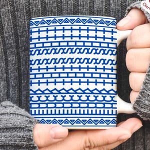 May include: White ceramic mug with a blue geometric pattern. The design features rows of triangles, lines, and wave-like shapes. The mug is held by a person wearing a gray sweater.