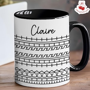 May include: A white ceramic mug with a black interior and handle. The mug features the name "Claire" in cursive, above a series of black geometric patterns. The mug is on a wooden surface.