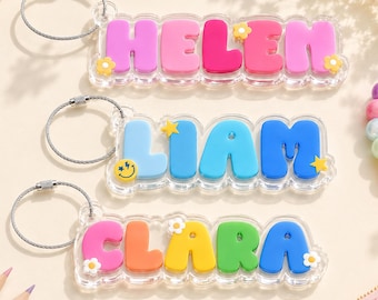 May include: Three clear acrylic keychains with metal wire loops. The top keychain features the name HELEN in pink letters with yellow flower accents. The middle keychain displays LIAM in blue letters with yellow stars and a smiley face. The bottom keychain shows CLARA in pink, orange, yellow, green, and blue letters with white flower accents.