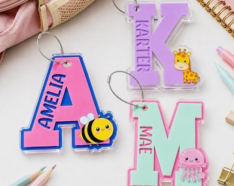 May include: Three acrylic letter keychains with metal rings. One pink 'A' with a bee and the name 'AMELIA', one purple 'K' with a giraffe and the name 'KARTER', and one mint green 'M' with a jellyfish and the name 'MAE' printed on the surface.