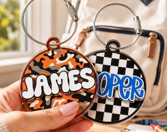 May include: Two circular acrylic bag tags attached to silver wire loops. One tag features an orange, black, and gray camouflage pattern with the white text JAMES. The other tag has a black and white checkerboard pattern with the blue text OPER.