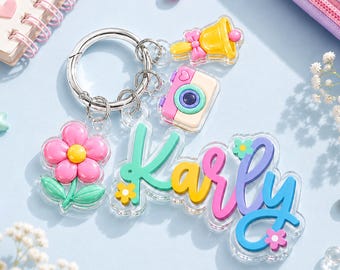 May include: A silver metal keychain featuring a clear acrylic nameplate with the name Karly written in colorful script. The keychain includes charms of a pink flower, a yellow bell with a pink bow, and a pastel-colored camera, all attached to a ring.