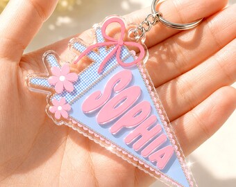 May include: A triangular acrylic keychain featuring a light blue background with a white gingham pattern. The name SOPHIA is written in large, raised pink letters. The design includes a pink bow at the top and two pink flowers on the left side.