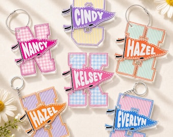 May include: Six acrylic letter keychains featuring various pastel gingham and striped patterns. Each letter is adorned with a colorful pennant flag displaying the names Nancy, Cindy, Hazel, Kelsey, Hazel, and Everlyn in white capital letters.