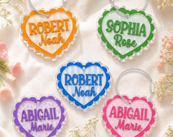 May include: Five heart-shaped acrylic luggage tags with scalloped edges and metal cable loops. Each tag features a unique background pattern and custom names in raised lettering: Robert Noah in orange, Sophia Rose in green, Robert Noah in blue, Abigail Marie in purple, and Abigail Marie in pink.