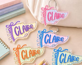 May include: Five acrylic pennant-shaped keychains featuring the name CLAIRE in various colors. Each keychain has a gingham pattern background, a ribbon-style border, and a metal ring. The colors include pink, purple, blue, orange, and green.