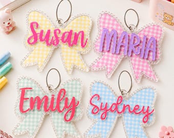 May include: Four acrylic bow-shaped ornaments featuring a gingham pattern in yellow, pink, green, and blue. Each ornament has a metal wire loop and a raised, script-style name tag in a contrasting color, displaying the names Susan, Maria, Emily, and Sydney.
