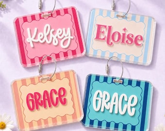 May include: Four rectangular acrylic luggage tags with rounded corners and metal cable loops. Each tag features a scalloped central frame over a striped background. The tags display the names Kelsey, Eloise, and Grace in various fonts and colors.