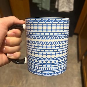 Puede incluir: Taza de cer&aacute;mica blanca con un patr&oacute;n geom&eacute;trico azul y un asa negra. La taza presenta un dise&ntilde;o repetitivo de l&iacute;neas, zigzags y tri&aacute;ngulos. La taza est&aacute; sujeta por una mano.