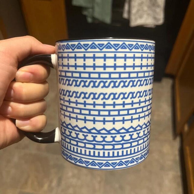 Puede incluir: Taza de cer&aacute;mica blanca con un patr&oacute;n geom&eacute;trico azul y un asa negra. La taza presenta un dise&ntilde;o repetitivo de l&iacute;neas, zigzags y tri&aacute;ngulos. La taza est&aacute; sujeta por una mano.