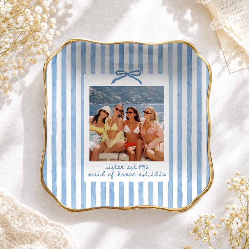 Puede incluir: Un plato decorativo cuadrado de cer&aacute;mica con bordes ondulados y un borde dorado. El plato presenta un patr&oacute;n de rayas de acuarela azul y blanco y una impresi&oacute;n fotogr&aacute;fica rectangular central de tres personas en traje de ba&ntilde;o, con el texto "sister est. 1996" y "maid of honor est. 2026" debajo de la imagen.
