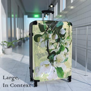 May include: A large rolling suitcase featuring a hard shell with a printed design of white apple blossoms and green leaves on a pale yellow background. The luggage has a black frame, a retractable handle, and four black wheels.