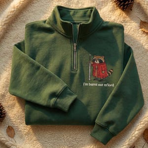 Może przedstawiać: Ciemnozielona bluza z zamkiem błyskawicznym do połowy szyi. Przedstawia rysunek szopa w czerwonym płaszczu trzymającego miecz, z napisem "I'm burnt out m'lord". Ze ściągaczem przy kołnierzu i mankietach.