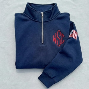 May include: Navy blue quarter-zip sweatshirt with a red embroidered monogram and an American flag patch on the sleeve. The sweatshirt has a ribbed collar and cuffs.