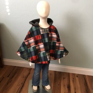 May include: A plaid hooded poncho with a patchwork design in red, green, black, and white. The poncho is on a mannequin wearing blue jeans.