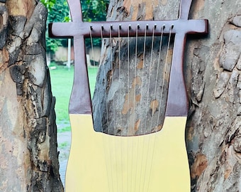 May include: A cream-coloured lyre with a wooden frame and strings. The instrument has a curved base and a horizontal bar at the top. The frame is a dark brown colour, and the strings are silver. The lyre is hanging on a tree.