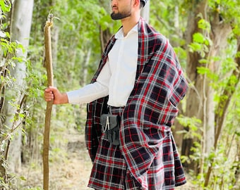 May include: A person wearing a traditional Scottish outfit, including a black and red plaid kilt, matching shawl, and black beret. The outfit is paired with a white shirt, cream-colored socks, and black boots. The person is holding a wooden walking stick.