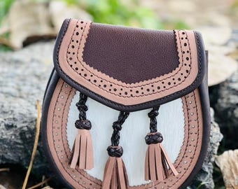 Op de afbeelding: Een bruine leren tas met een wit middenpaneel en decoratieve stiksels. De tas heeft een flap met een geperforeerd ontwerp en drie bruine kwastjes. De tas ligt op een rots.
