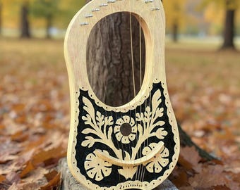 May include: A wooden lyre with a light-coloured frame and a black, floral-patterned soundboard. The instrument has strings and is resting on a concrete block outdoors, surrounded by autumn leaves. The lyre is approximately 51 cm tall.