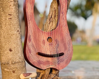 May include: A reddish-brown lyre, a stringed musical instrument, is displayed outdoors. The instrument has a rounded body with decorative carvings and a sound hole. A tuning tool rests below the lyre. The lyre is approximately 20 inches in height.