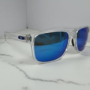 May include: Clear-framed sunglasses with blue mirrored lenses. The sunglasses have a rectangular shape and a transparent frame. The lenses reflect a bright blue color. The sunglasses are displayed on a white marble surface.