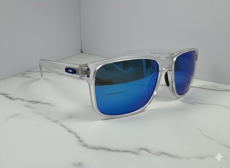 May include: Clear-framed sunglasses with blue mirrored lenses. The sunglasses have a rectangular shape and a transparent frame. The lenses reflect a bright blue color. The sunglasses are displayed on a white marble surface.