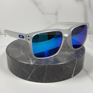 Puede incluir: Gafas de sol con montura plateada y lentes azules espejadas. Las gafas se exhiben sobre un pedestal cil&iacute;ndrico de m&aacute;rmol gris sobre un fondo de m&aacute;rmol blanco. Las gafas tienen forma rectangular y un peque&ntilde;o logotipo azul en las patillas.