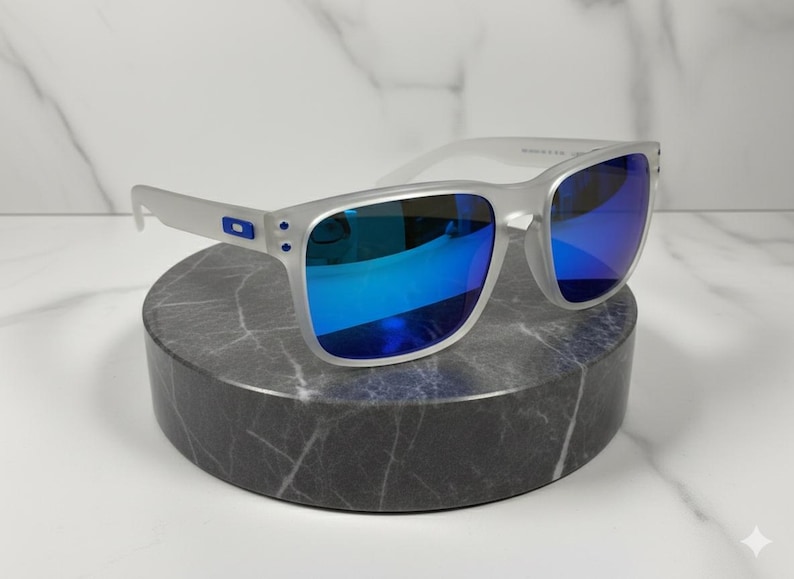 Puede incluir: Gafas de sol con montura plateada y lentes azules espejadas. Las gafas se exhiben sobre un pedestal cil&iacute;ndrico de m&aacute;rmol gris sobre un fondo de m&aacute;rmol blanco. Las gafas tienen forma rectangular y un peque&ntilde;o logotipo azul en las patillas.