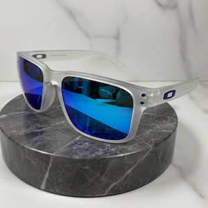Puede incluir: Gafas de sol con montura transparente y lentes azules espejadas. Las gafas se exhiben sobre un pedestal de piedra circular gris oscuro. Las lentes reflejan un tono azul brillante, y la montura tiene una calidad sutil y transl&uacute;cida.