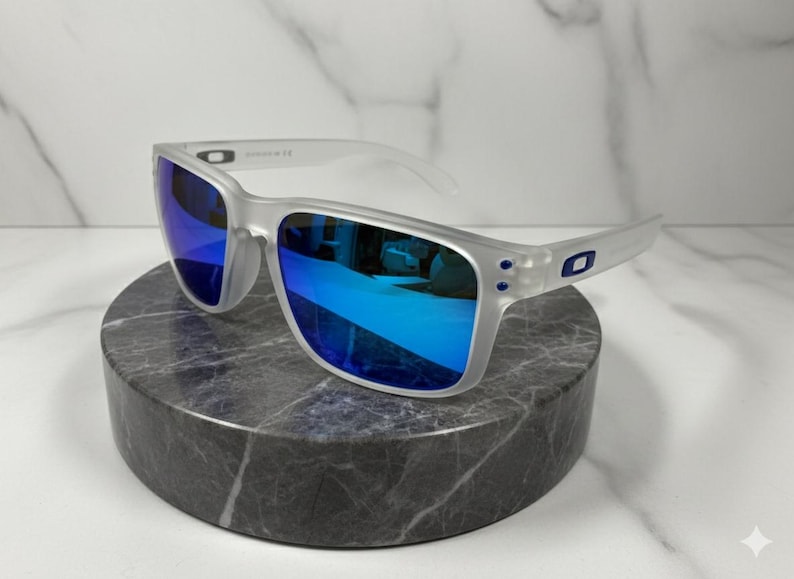 Puede incluir: Gafas de sol con montura transparente y lentes azules espejadas. Las gafas se exhiben sobre un pedestal de piedra circular gris oscuro. Las lentes reflejan un tono azul brillante, y la montura tiene una calidad sutil y transl&uacute;cida.