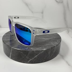 Puede incluir: Gafas de sol con montura transparente y lentes azules espejadas. Las gafas de sol tienen forma rectangular y est&aacute;n apoyadas sobre una piedra circular gris oscuro. El logotipo de Oakley es visible en las patillas.