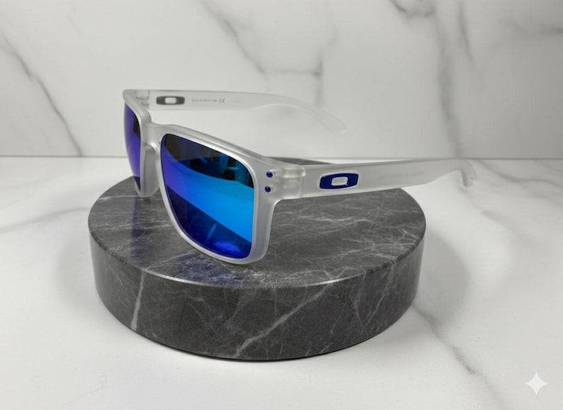 Puede incluir: Gafas de sol con montura transparente y lentes azules espejadas. Las gafas de sol tienen forma rectangular y est&aacute;n apoyadas sobre una piedra circular gris oscuro. El logotipo de Oakley es visible en las patillas.