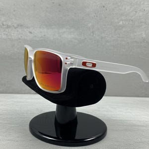 Puede incluir: Gafas de sol con montura blanca y lentes rojas. Las gafas se exhiben en un soporte negro. El logotipo de Oakley es visible en la patilla.