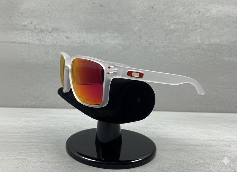 Puede incluir: Gafas de sol con montura blanca y lentes rojas. Las gafas se exhiben en un soporte negro. El logotipo de Oakley es visible en la patilla.