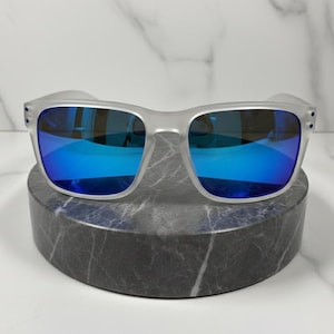 Puede incluir: Unas gafas de sol gris claro con lentes azules espejadas. Las gafas de sol se exhiben sobre un pedestal de m&aacute;rmol gris oscuro sobre un fondo de m&aacute;rmol blanco. Las monturas tienen forma cuadrada.