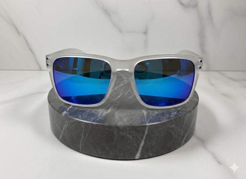 Puede incluir: Unas gafas de sol gris claro con lentes azules espejadas. Las gafas de sol se exhiben sobre un pedestal de m&aacute;rmol gris oscuro sobre un fondo de m&aacute;rmol blanco. Las monturas tienen forma cuadrada.