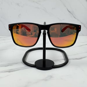 May include: Black sunglasses with orange-tinted lenses are displayed on a black metal stand. The sunglasses have a square frame with red accents on the temples. The lenses reflect the surrounding environment.