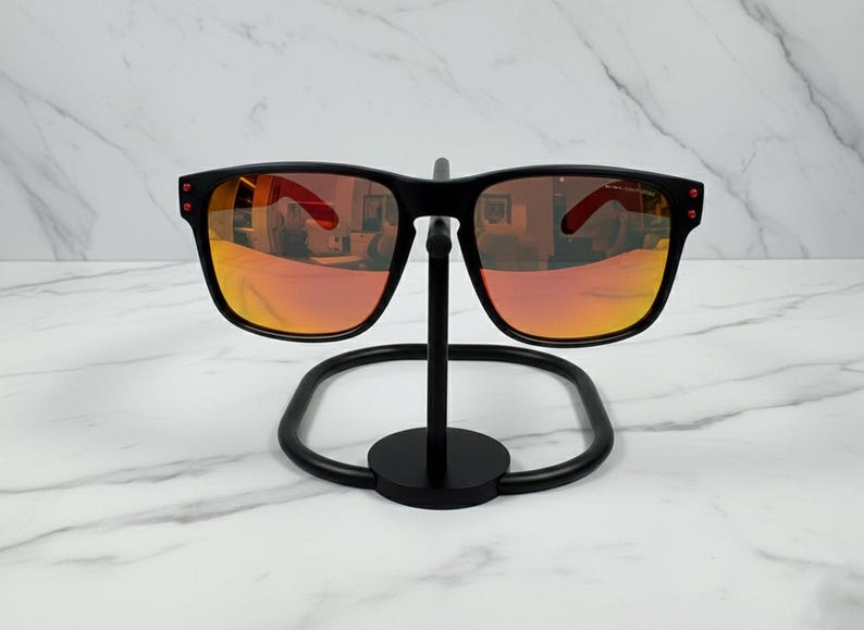 May include: Black sunglasses with orange-tinted lenses are displayed on a black metal stand. The sunglasses have a square frame with red accents on the temples. The lenses reflect the surrounding environment.