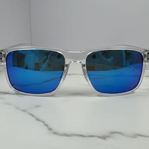 May include: Clear-framed sunglasses with vibrant blue mirrored lenses. The square-shaped frames are transparent, offering a modern aesthetic. The sunglasses are displayed on a white marble surface, highlighting their sleek design.
