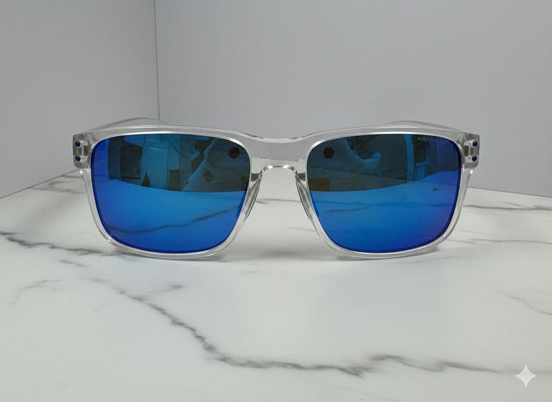 May include: Clear-framed sunglasses with vibrant blue mirrored lenses. The square-shaped frames are transparent, offering a modern aesthetic. The sunglasses are displayed on a white marble surface, highlighting their sleek design.
