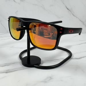 May include: Black sunglasses with orange-tinted lenses are displayed on a black metal stand. The sunglasses have a rectangular frame with red accents on the temples. The stand has a circular base and a curved design.