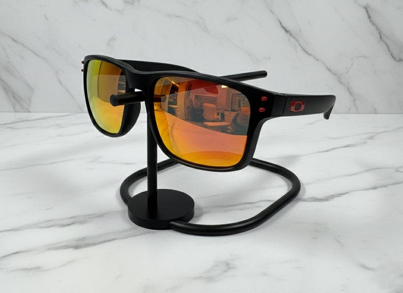 May include: Black sunglasses with orange-tinted lenses are displayed on a black metal stand. The sunglasses have a rectangular frame with red accents on the temples. The stand has a circular base and a curved design.
