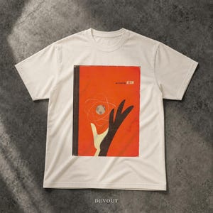 Può includere: Una t-shirt a maniche corte color bianco sporco con una stampa grafica rettangolare sul petto. La stampa raffigura una copertina di libro arancione stilizzata con una mano nera che si protende verso un diagramma atomico bianco e il testo "we found the ATOM".