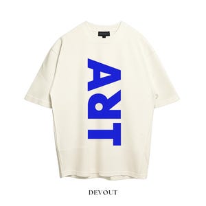 May include: An off-white short-sleeve t-shirt featuring the word ART printed vertically in bold blue capital letters across the center. The garment has a crew neckline and a black interior neck label with the brand name DEVOUT printed in white.
