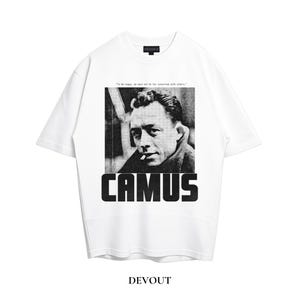 May include: A white short-sleeved t-shirt featuring a black and white photographic print of Albert Camus with a cigarette. Above the portrait is the text "To be happy, we must not be too concerned with others." and below is the name "CAMUS" in bold.