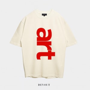 以下が含まれることがあります： クリーム色のTシャツ。赤色の太字で「art」の文字が縦にプリントされています。Tシャツは、クラシックなクルーネックと半袖です。「DEVOUT」のブランド名が下部に黒色でプリントされています。