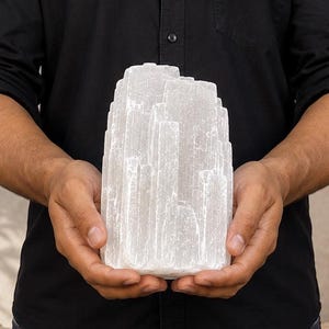 Può includere: Una torre di cristallo di selenite bianca traslucida con un bordo superiore naturale e frastagliato e una base rettangolare. Il cristallo presenta striature verticali e una consistenza fibrosa e cristallina su tutta la superficie, tenuto con entrambe le mani.