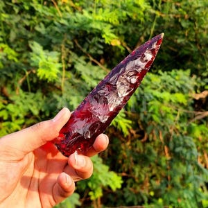 May include: A hand holds a translucent, deep red arrowhead or blade. The object has a sharp point and a textured surface, catching the light. The background is a blurred view of green foliage.