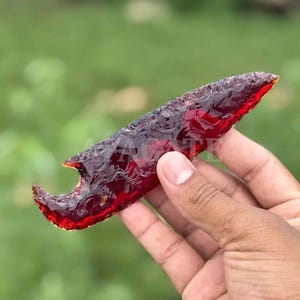 Puede incluir: Una mano sostiene una punta de flecha translúcida de color rojo oscuro. La punta de flecha tiene una base curva y una punta afilada. El fondo es un verde borroso.